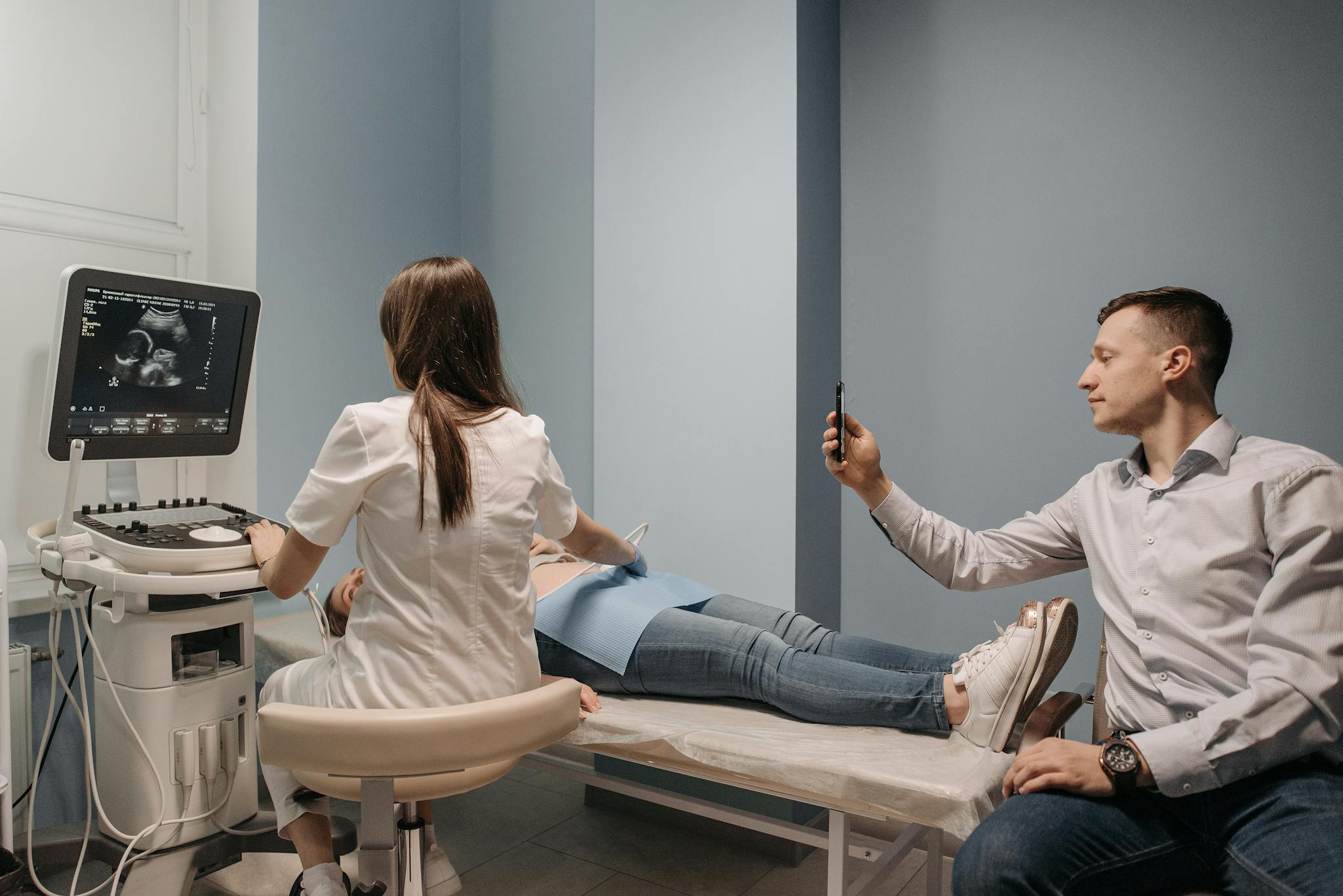 A doctor performs an ultrasound on a pregnant woman, while a man takes a photo in a clinical environment.