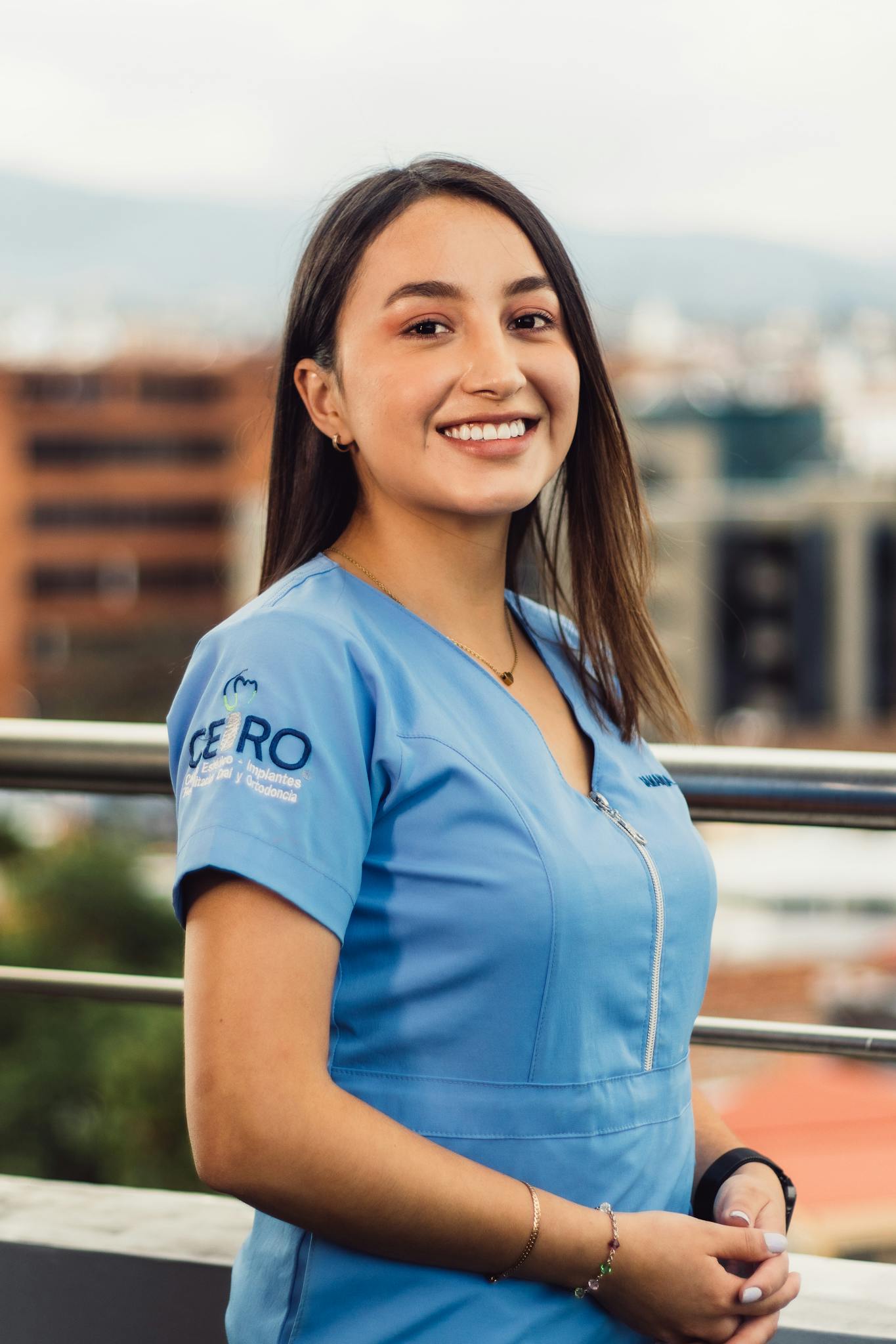 Young female nurse smiling outdoors with urban background, wearing blue uniform.